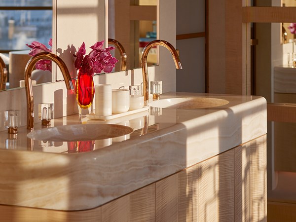 Light reflecting on wall behind sink and gold taps with purple flower on the side in a vase